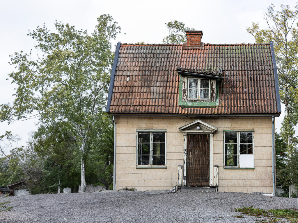 An aged house stands in a natural setting. The house has beige walls consisting of asbestos and a brown-tiled roof. There’s an attic window with green trimmings, and two white windows flank a worn wooden door. The ground is covered in gravel, and tall trees surround the house under a cloudy sky. A renovation team would be very well advised if they would carry out a Raman Asbestos fiber analysis.