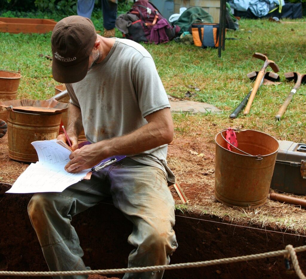 FTIR in archaeological excavations. The image depicts an individual wearing a cap, sitting outdoors at an archaeological site. They are writing on a piece of paper, surrounded by excavation tools like shovels and buckets. 