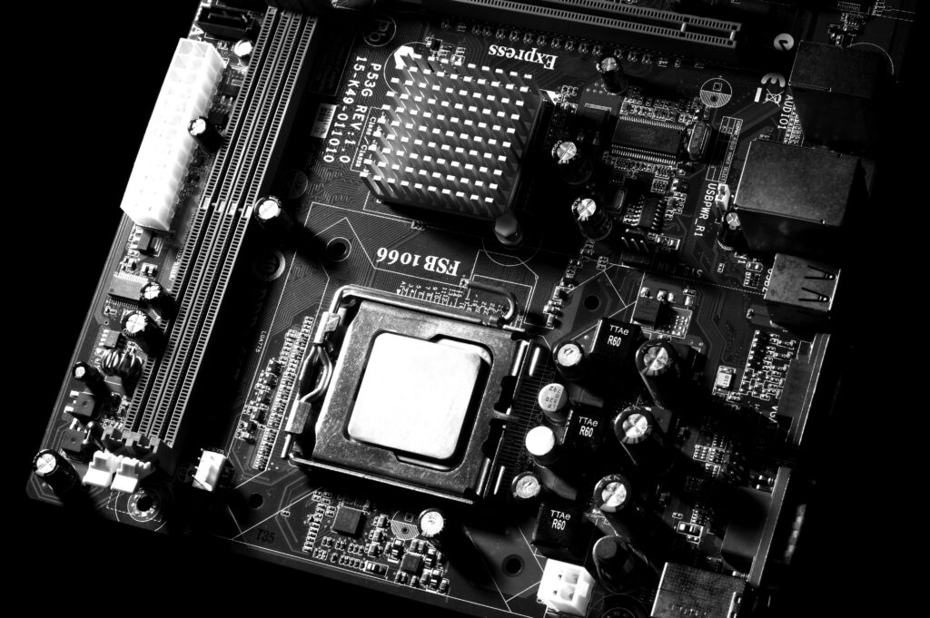 Black and white image of a motherboard. The image shows a flat, rectangular board filled with small, intricate components, including cylindrical shapes (capacitors), a large square area (CPU socket), and various tiny, rectangular chips, all connected by thin lines (circuit traces).



