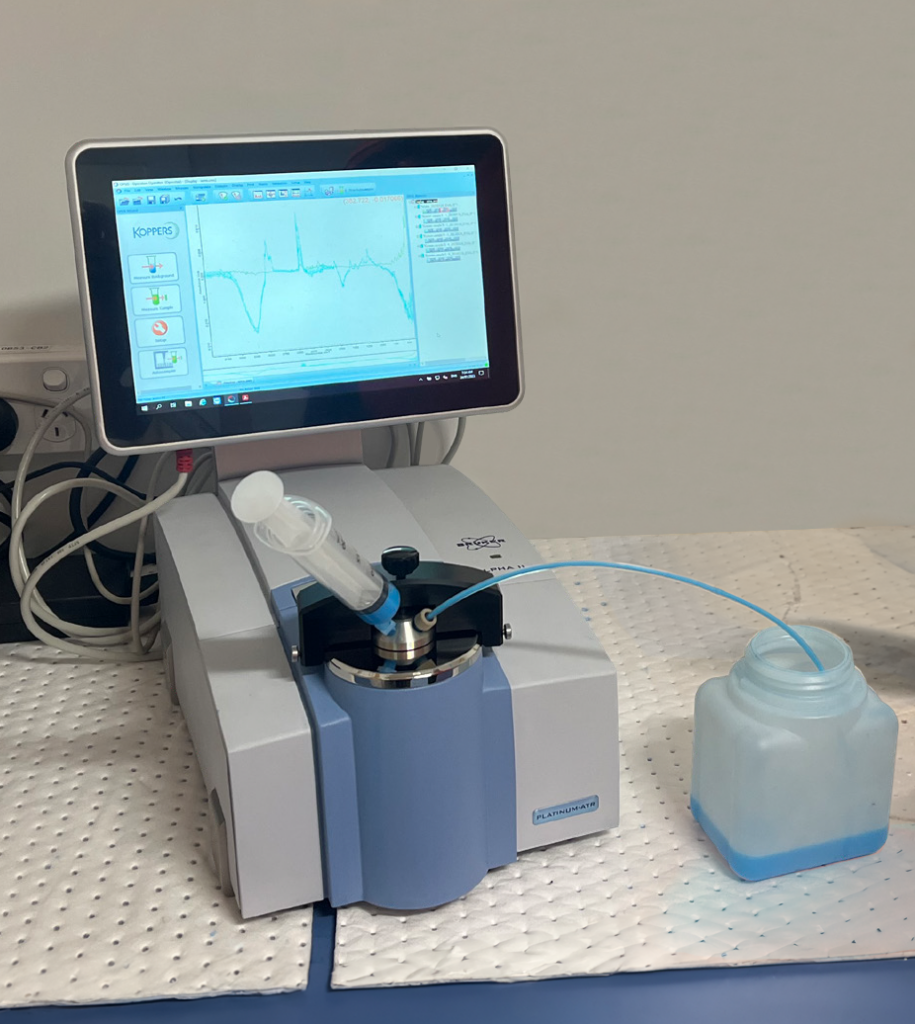 FTIR device in blue and white. A syringe is inside of the ATR. This device is used for chemical retention analysis where FTIR aids wood preservation.